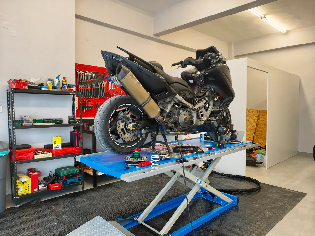 A black scooter on the workshop's blue scissor lift. Behind it, the red tool wall.
