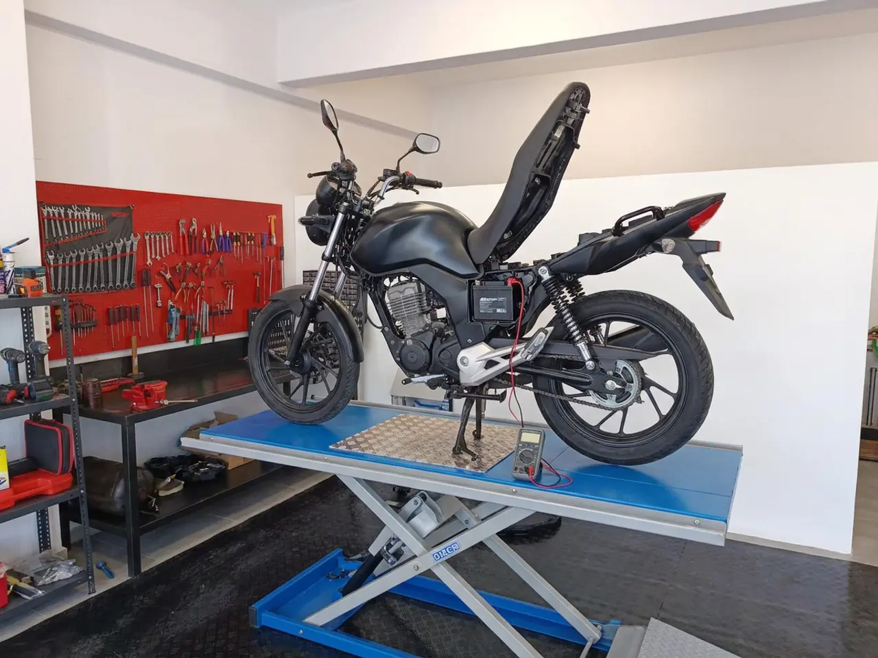 A battery being tested with a multimeter on a motorcycle with its seat open, on the blue lift.